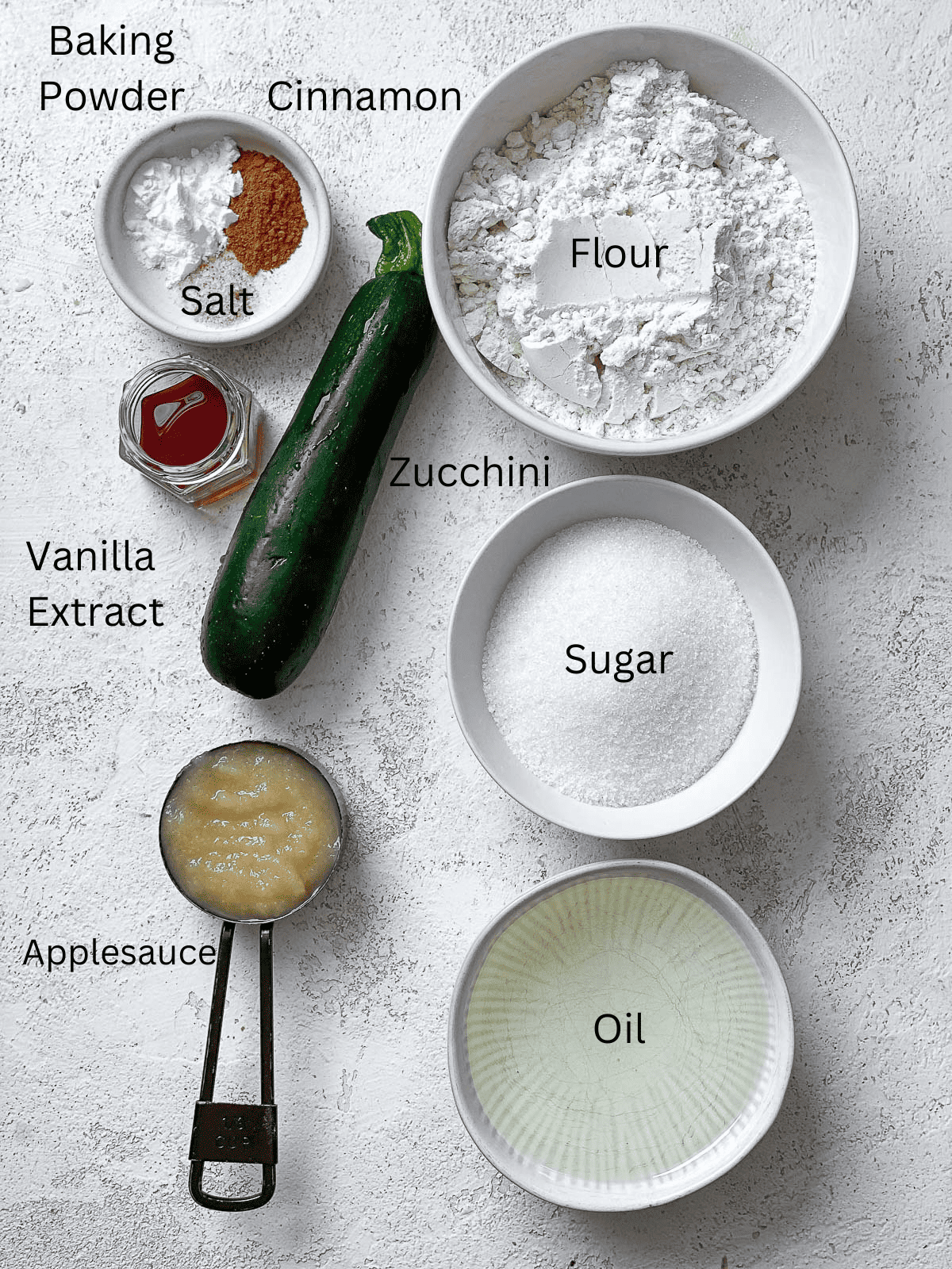 A photo of all the ingredients for Vegan Zucchini Bread place in small bowls sitting on textured white surface.