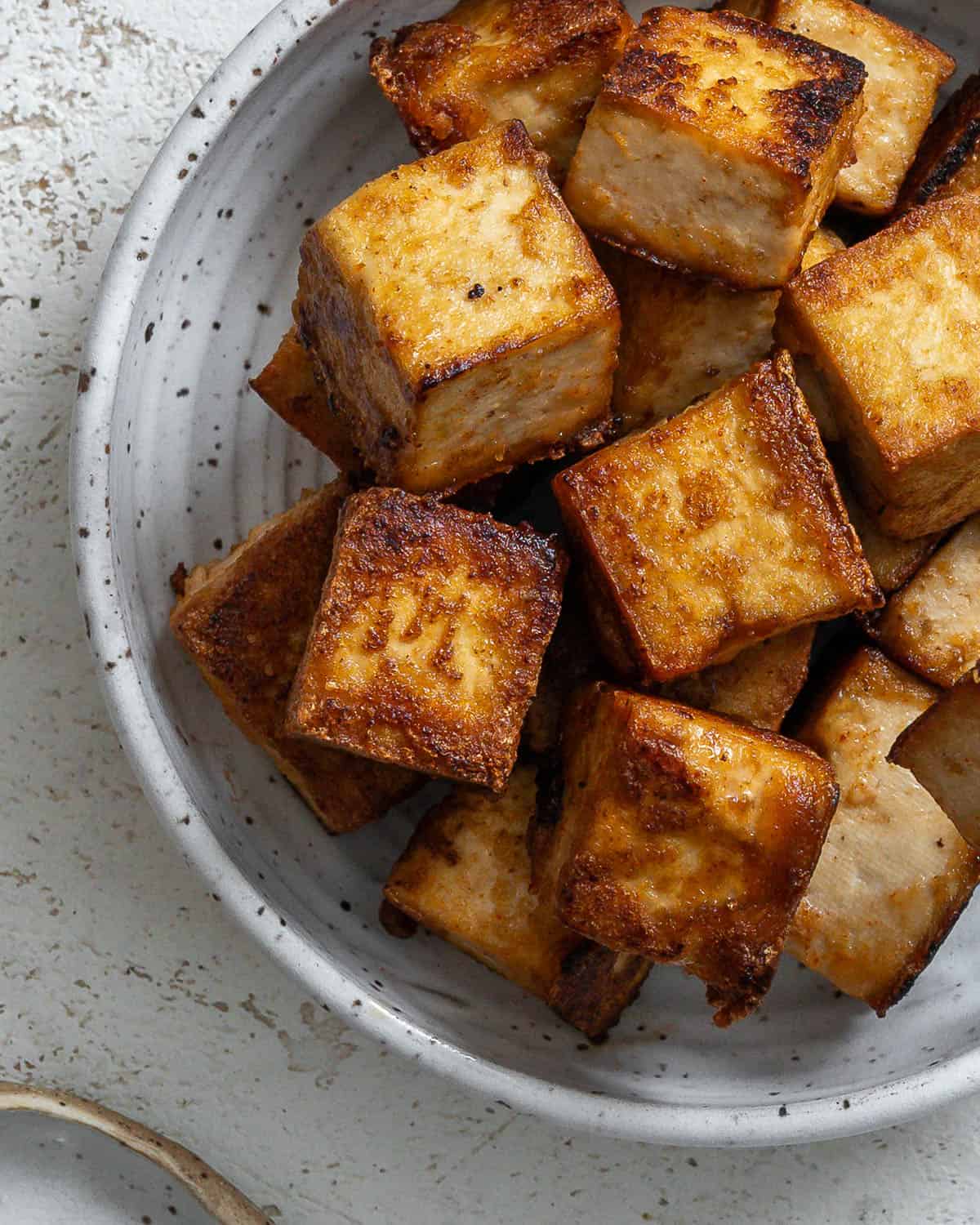 Pan fried tofu in a white speckled bowl on a textured white surface.