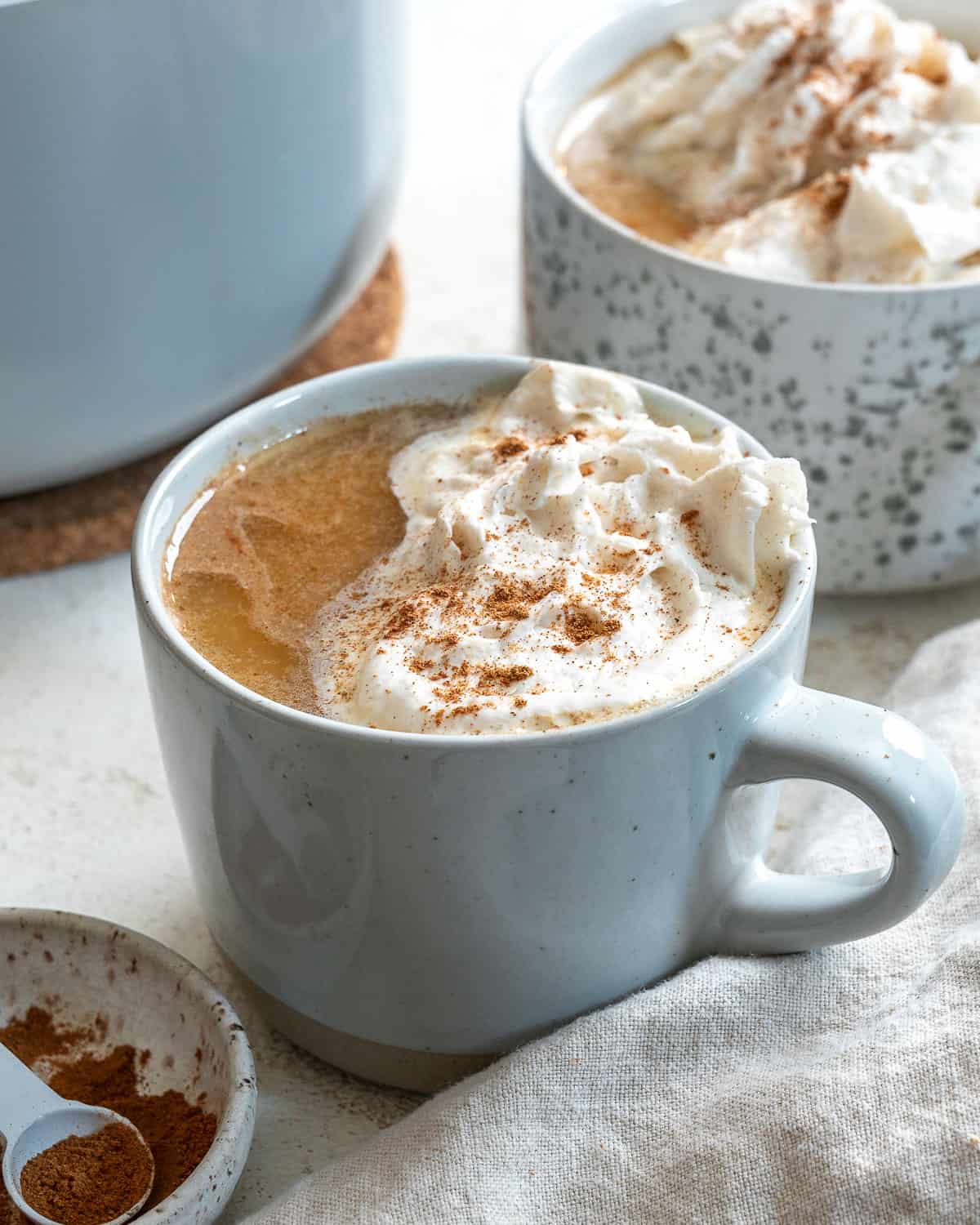 A finished vegan pumpkin spice latte topped with vegan whipped cream. On the top left corner, theres part of another glass with the same drink. Bottom right has a gray napkin. Bottom left has a small bowl of cinnamon.