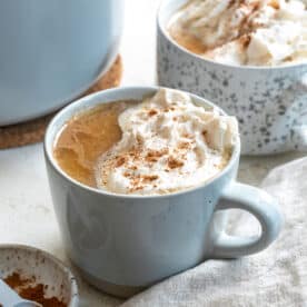 A finished vegan pumpkin spice latte topped with vegan whipped cream. On the top left corner, theres part of another glass with the same drink. Bottom right has a gray napkin. Bottom left has a small bowl of cinnamon.
