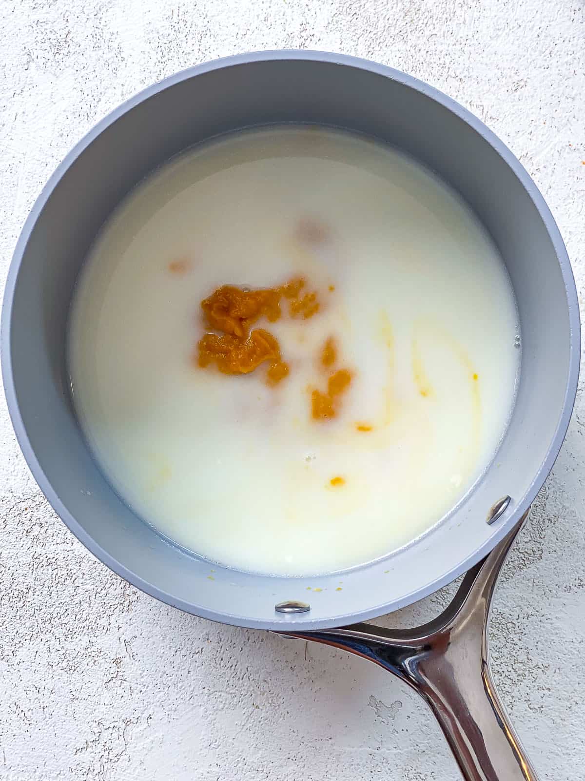 A pot with milk and pumpkin inside of it. The pot is gray with a silver handle and sitting on top of a textured white surface.