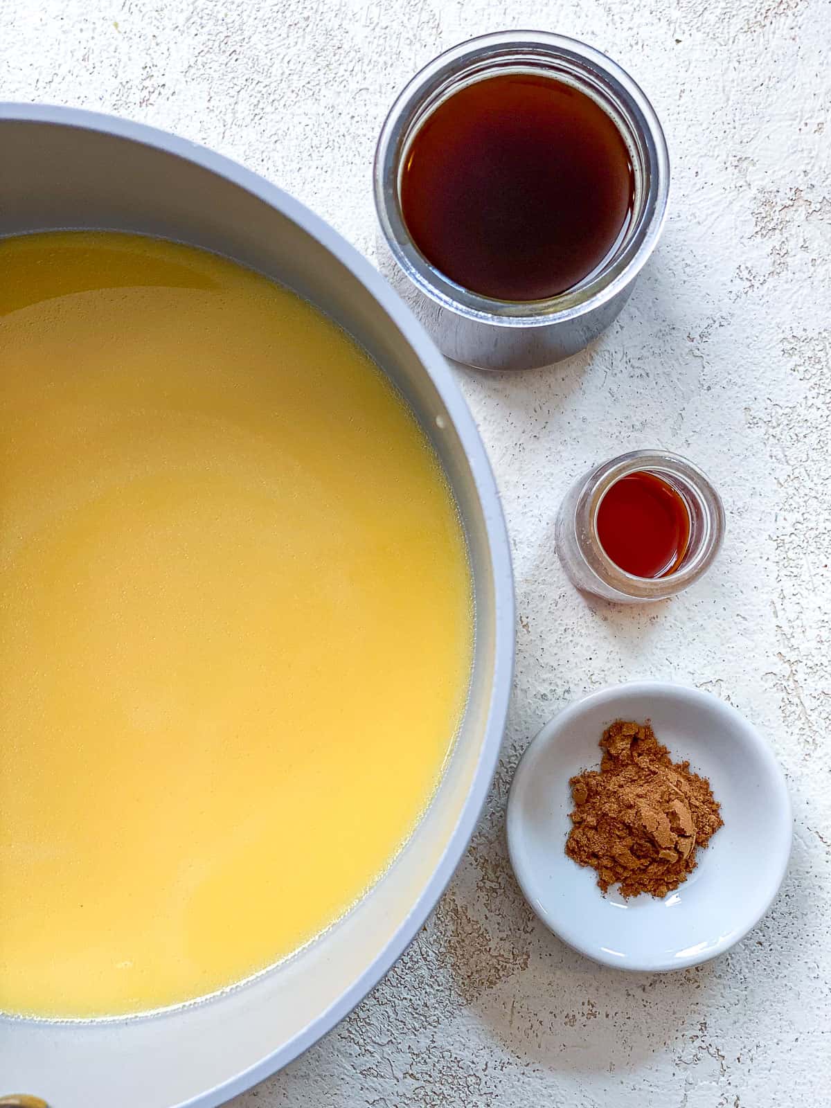 A pot with milk and pumpkin mixed inside of it. The pot is gray with a silver handle and sitting on top of a textured white surface. Beside the pot is a small bowl of sweetener, small bowl of vanilla, and a small bowl of pumpkin pie spice.