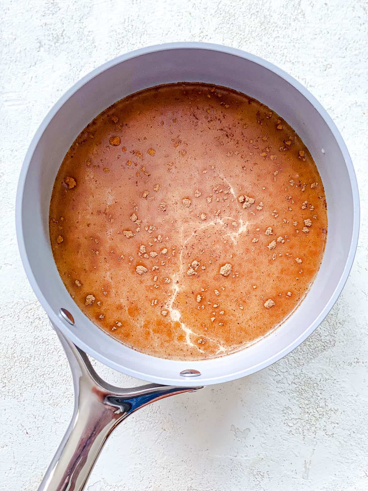 A pot with milk, pumpkin pie spice, and pumpkin mixed inside of it. The pot is gray with a silver handle and sitting on top of a textured white surface.