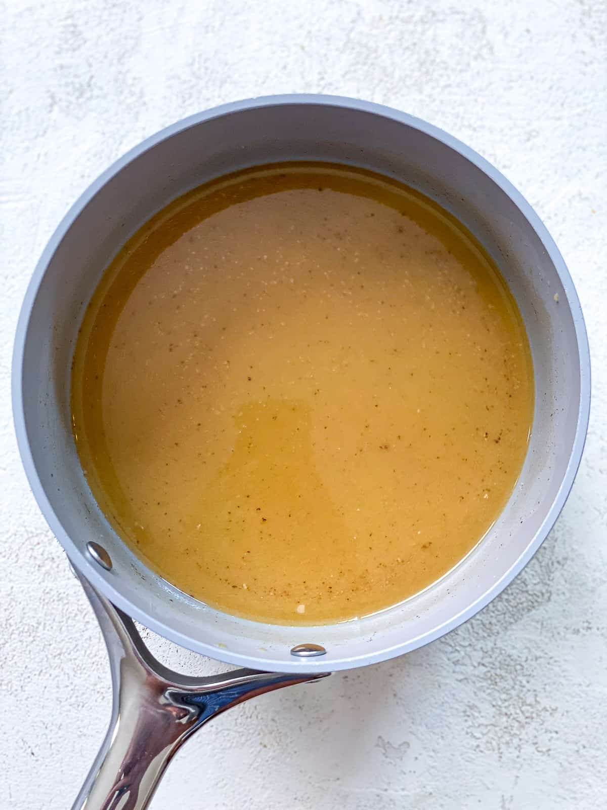 A pot with milk, pumpkin pie spice, vanilla, sweetener, and pumpkin inside of it. The pot is gray with a silver handle and sitting on top of a textured white surface.