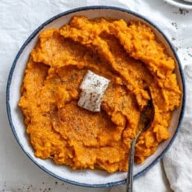 Cooked mashed vegan sweet potatoes with a slice of vegan butter on top. In a white bowl with a blue rim and a silver spoon on top.