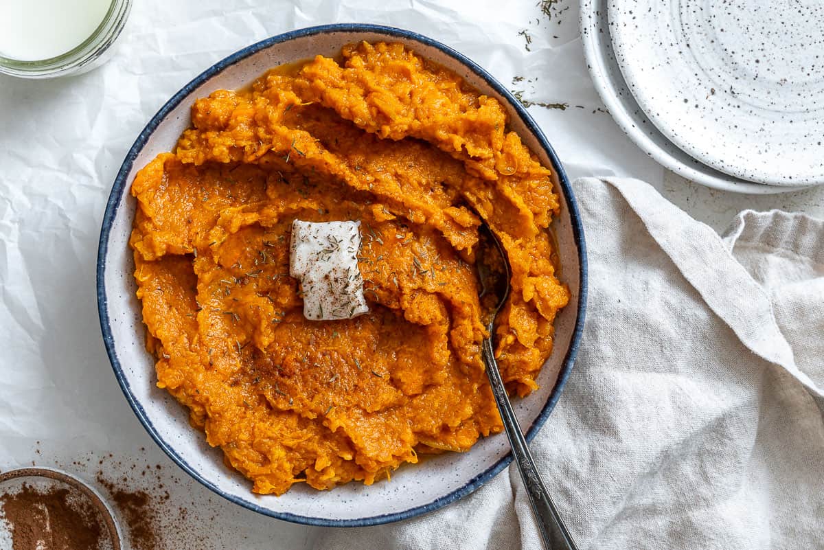 Cooked mashed vegan sweet potatoes with a slice of vegan butter on top. In a white bowl with a blue rim and a silver spoon on top. On a white crinkled paper with a white cloth napkin in the right bottom corner and cinnamon in the left corner,