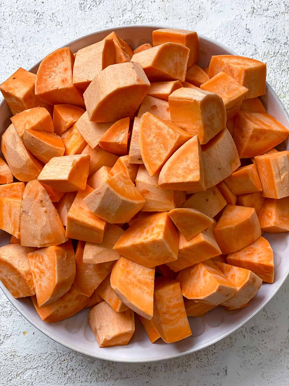 Bowl of raw chopped and peeled sweet potatoes in a white bowl on a textured white surface.