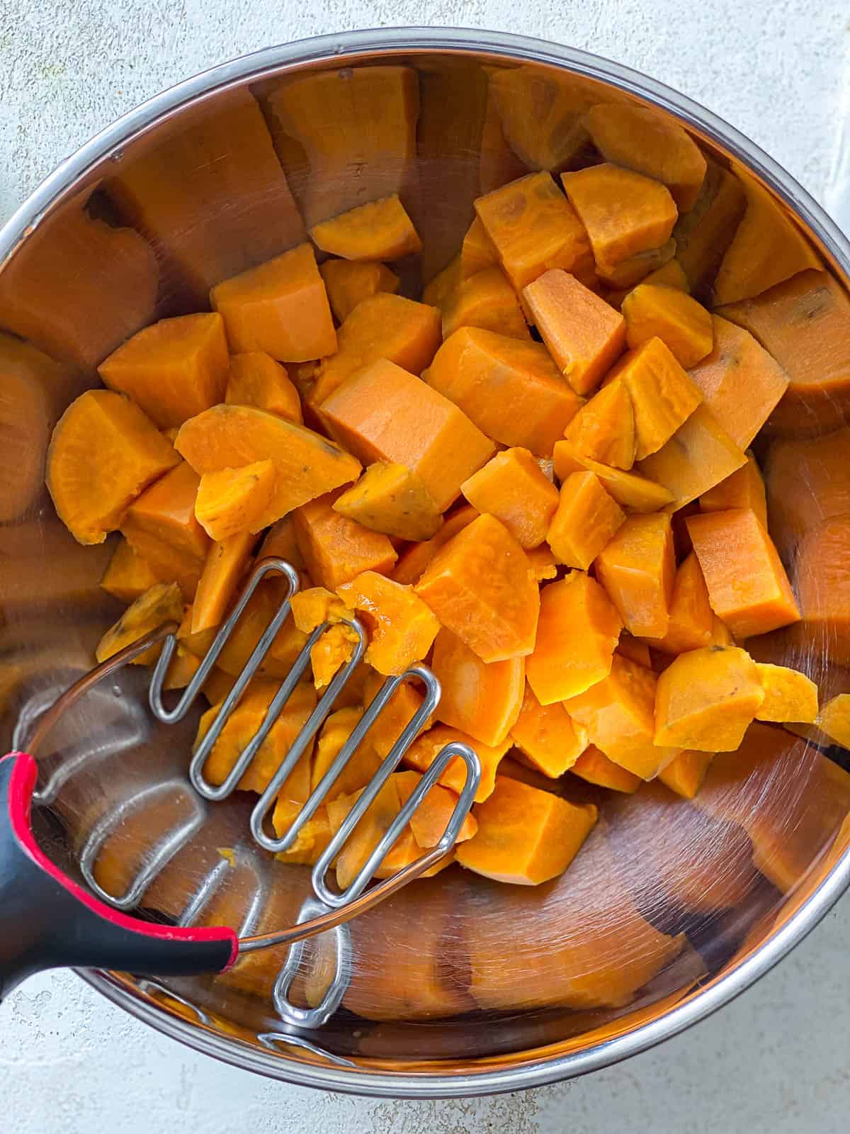 Cooked sweet potatoes in a silver bowl with a potato masher.