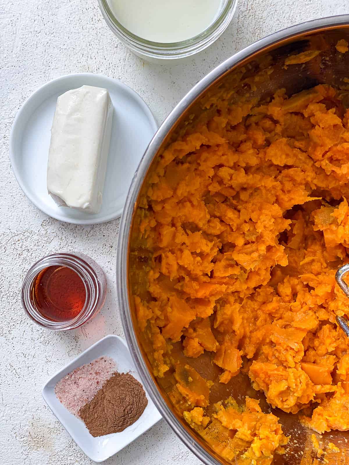 Mashed sweet potatoes in a silver bowl with vegan butter, plant milk, salt, cinnamon, and maple syrup on the left side of the bowl in small containers.