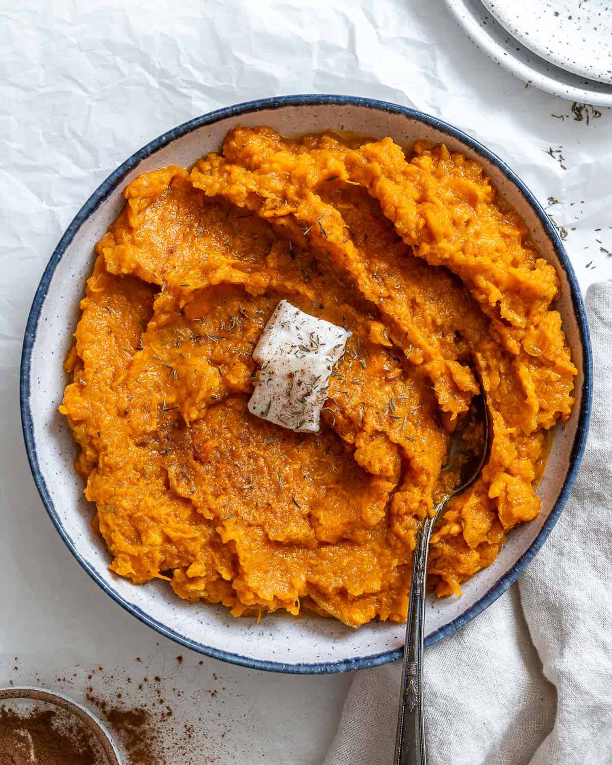 Cooked mashed vegan sweet potatoes with a slice of vegan butter on top. In a white bowl with a blue rim and a silver spoon on top. On a white crinkled paper with a white cloth napkin in the right bottom corner and cinnamon in the left corner,