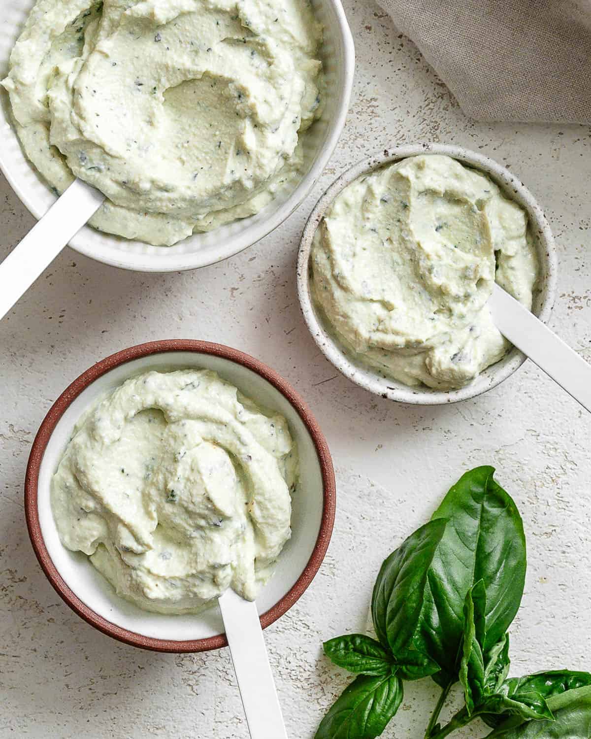 Three small bowls of tofu ricotta on white textured surface. Each bowl has a white spoon and there's a small bunch of fresh basil at the bottom right corner.
