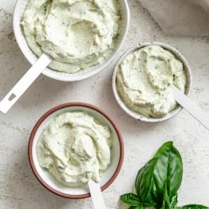 Three small bowls of tofu ricotta with white spoons in them with a fresh bunch of basil in the bottom right cornern.