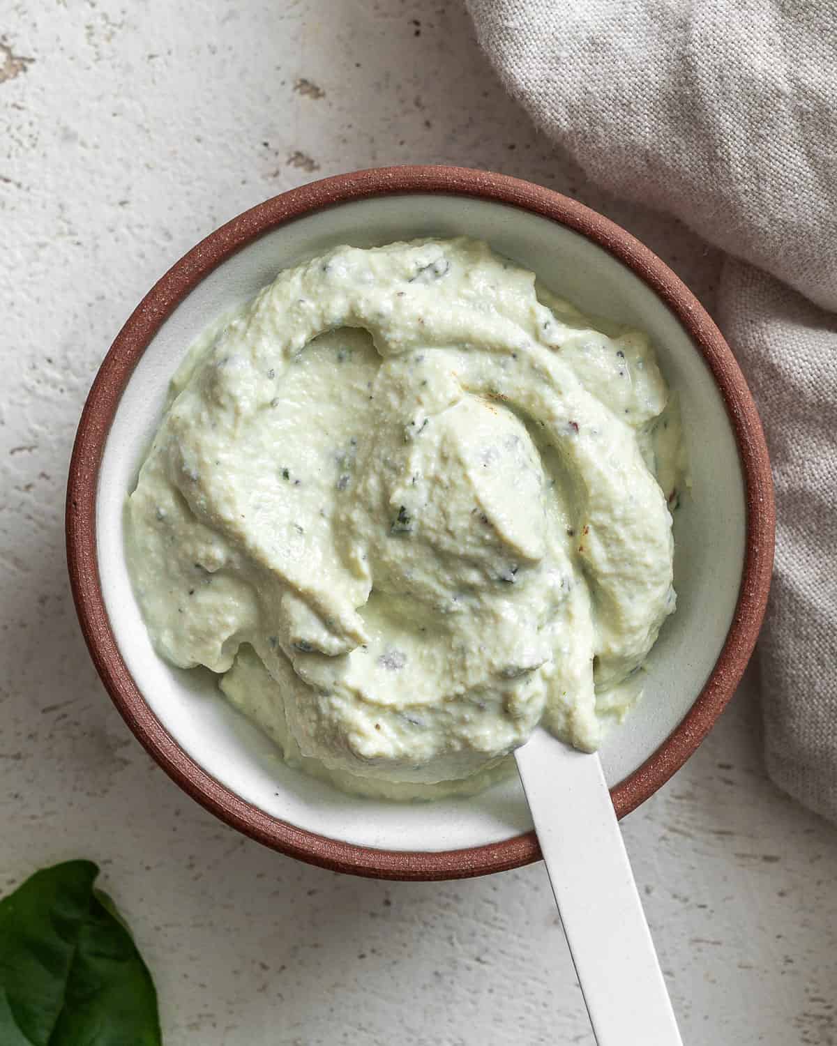 Bowl of tofu ricotta on a textured white surface with a white spoon in it and a leaf of basil in the bottom left corner.