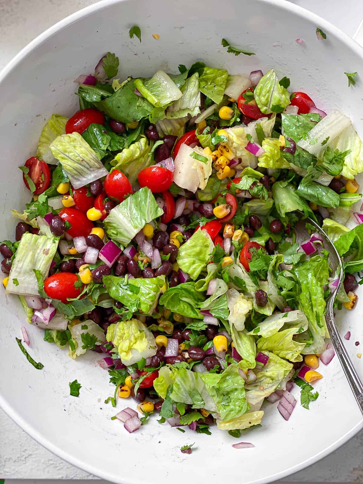 Large white bowl of chopped romaine lettuce, roasted corn, chopped cilantro, black beans, halved cherry tomatoes, and diced onions all mixed together.