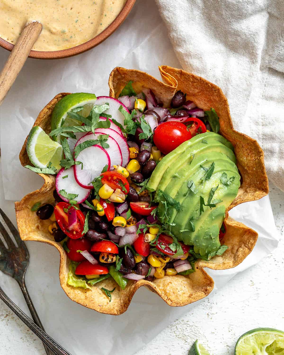 A tortilla bowl holding a taco salad that's topped with a sliced avocado, taco salad dressing, sliced radishes, and lime wedges. In the top left corner there's a small bowl of dressing with a wooden spoon in it. In the top right corner there's a gray napkin. In the bottom left corner there's a small bowl of chopped cilantro next to half of an avocado and sliced radishes. The background is white and textured.