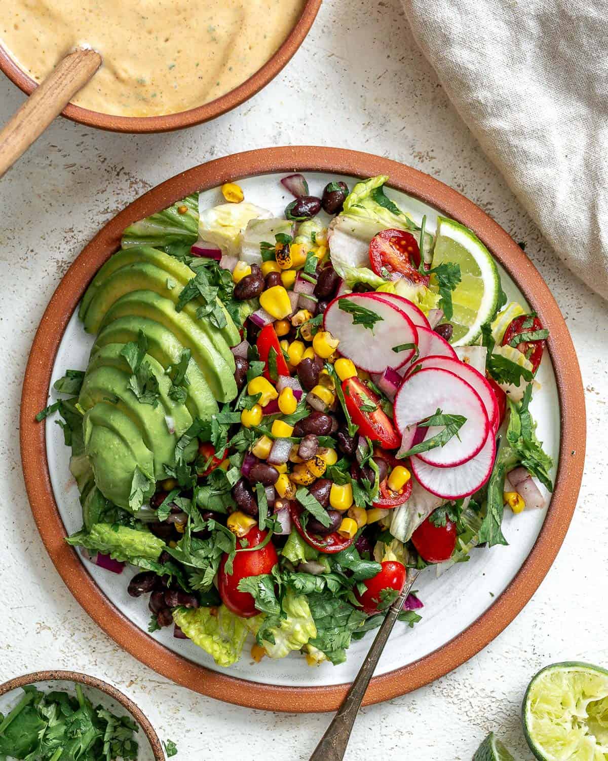 A taco salad that's topped with a sliced avocado, taco salad dressing, sliced radishes, and lime wedges. In the top left corner there's a small bowl of dressing with a wooden spoon in it. In the top right corner there's a gray napkin. In the bottom left corner there's a small bowl of chopped cilantro next to half of an avocado and sliced radishes. The background is white and textured.