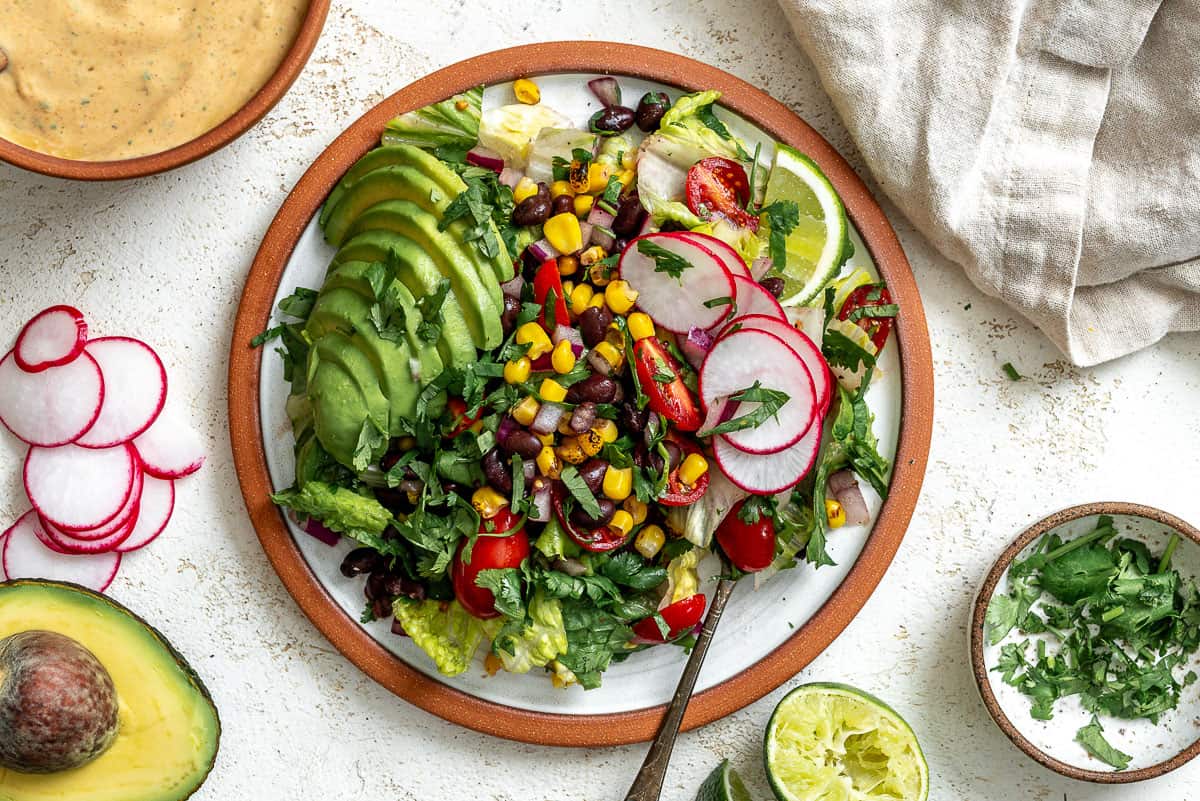 A taco salad that's topped with a sliced avocado, taco salad dressing, sliced radishes, and lime wedges. In the top left corner there's a small bowl of dressing with a wooden spoon in it. In the top right corner there's a gray napkin. In the bottom left corner there's a small bowl of chopped cilantro next to half of an avocado and sliced radishes. The background is white and textured.