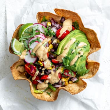 A tortilla bowl holding a taco salad that's topped with a sliced avocado, taco salad dressing, sliced radishes, and lime wedges