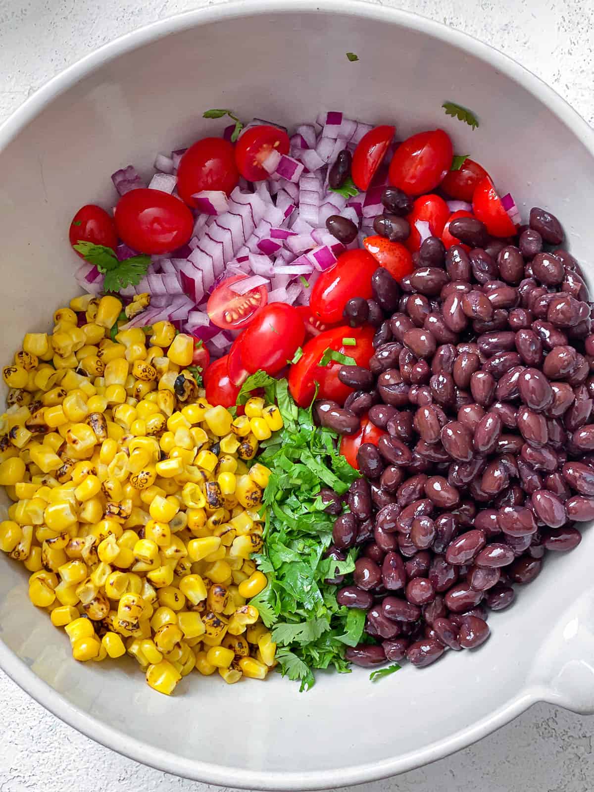 Large white bowl of roasted corn, chopped cilantro, black beans, halved cherry tomatoes, and diced onions.