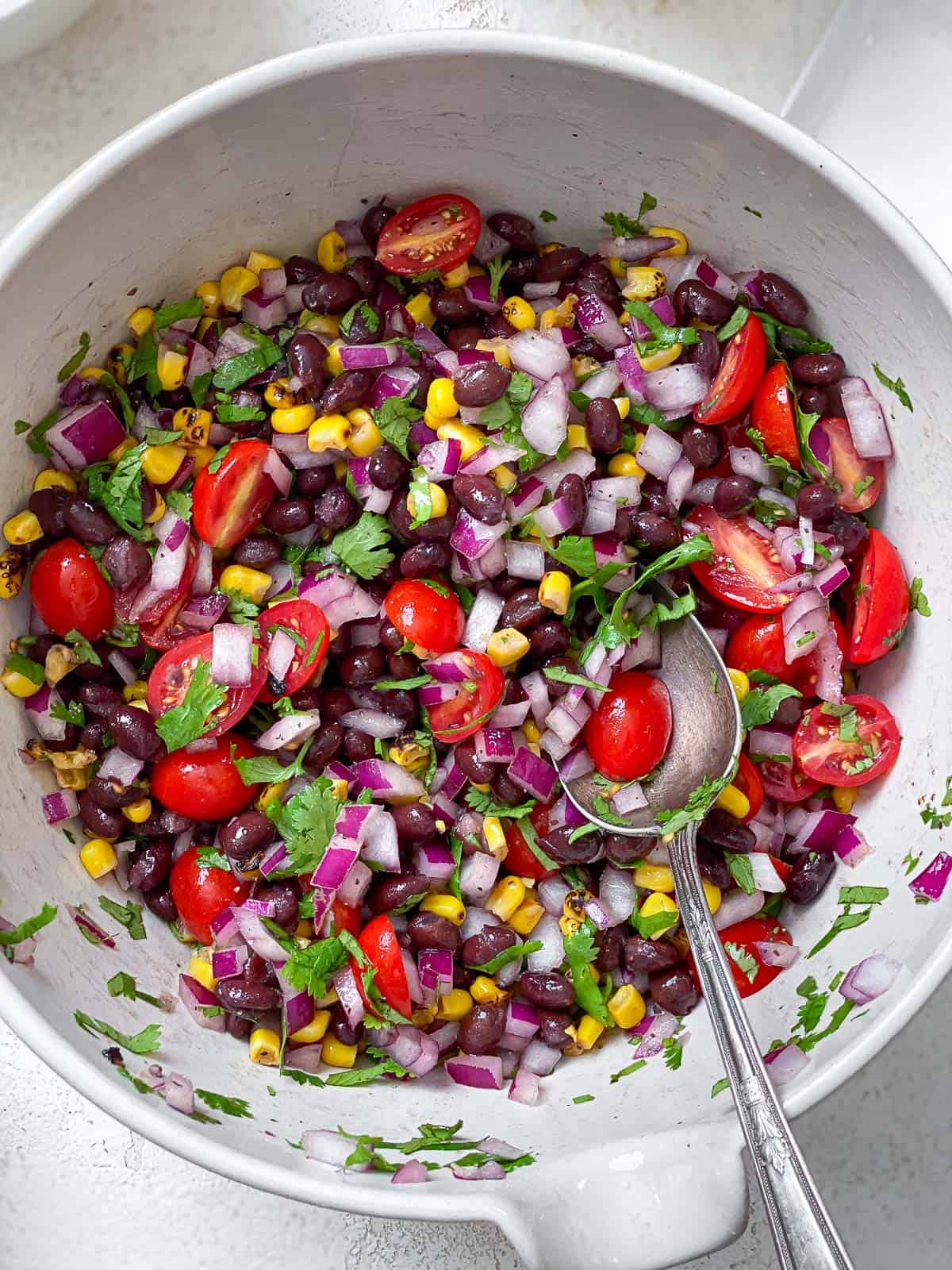 Large white bowl of roasted corn, chopped cilantro, black beans, halved cherry tomatoes, and diced onions all mixed together with a silver spoon on the bottom right corner of the bowl..