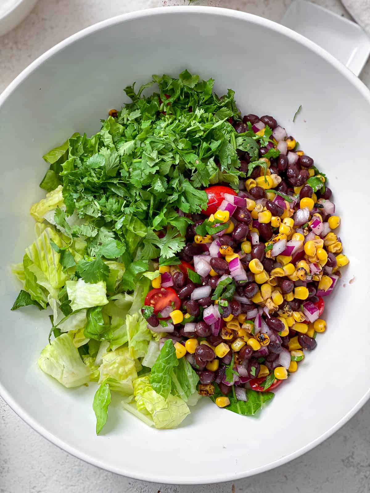 Large white bowl of chopped romaine lettuce, roasted corn, chopped cilantro, black beans, halved cherry tomatoes, and diced onions.