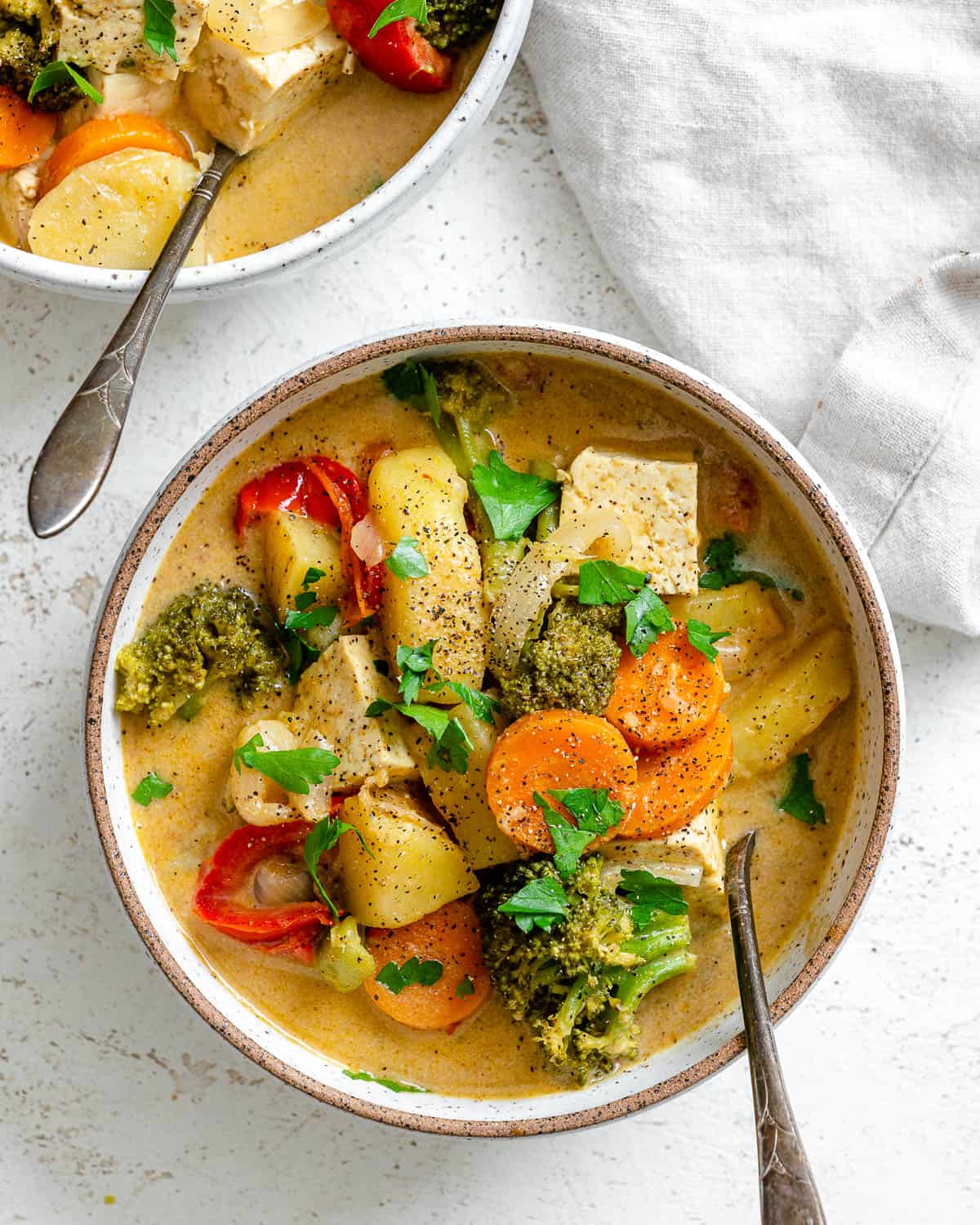 Bowl with Tofu Yellow Curry over a white textured surface with a spoon in it. There's also part of a bowl with curry in the top left corner.