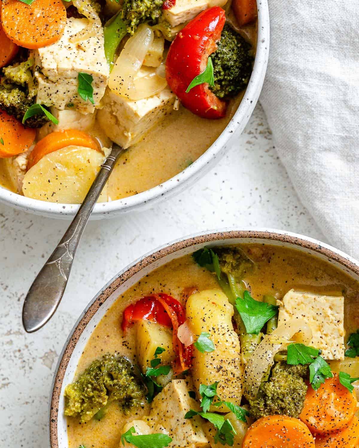 Bowl with Tofu Yellow Curry over a white textured surface with a spoon in it. There's also part of a bowl with curry in the top left corner.