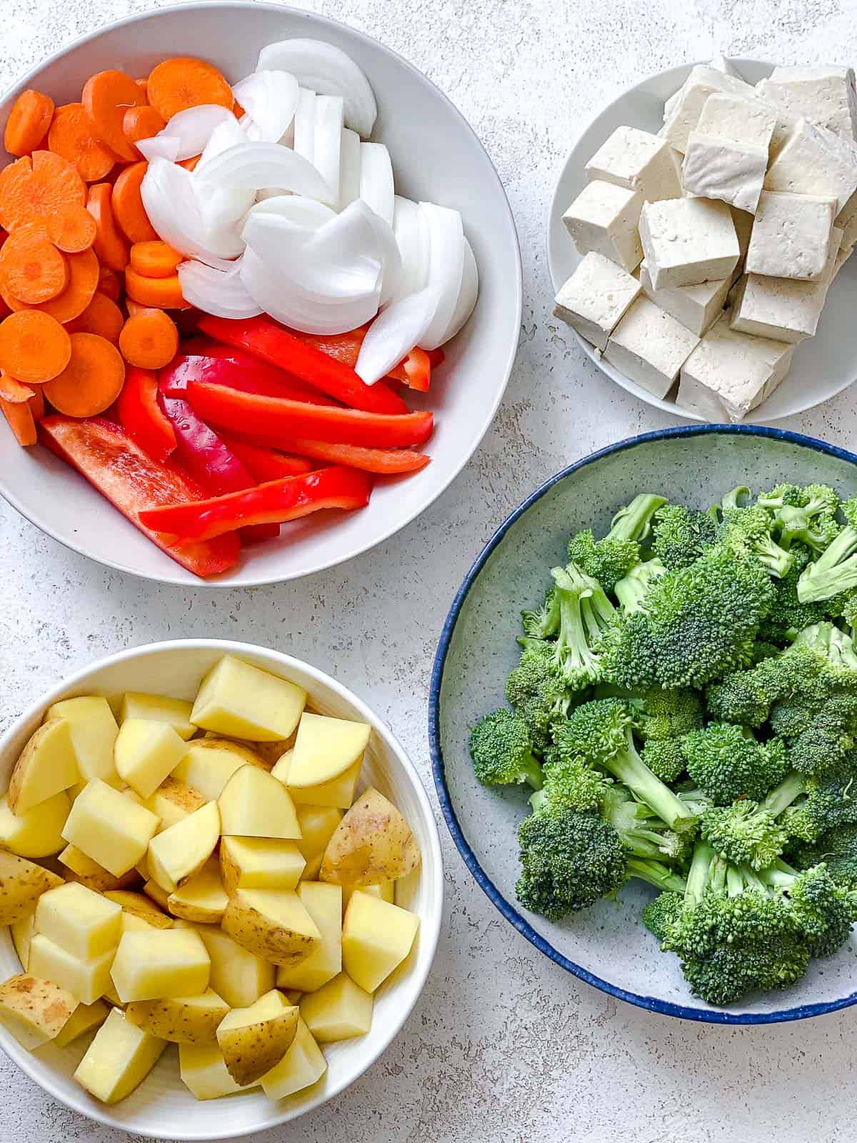 Bowl of chopped potatoes, bowl of broccoli florets, bowl of sliced red bell peppers, bowl of cubed tofu on a white textured surface.