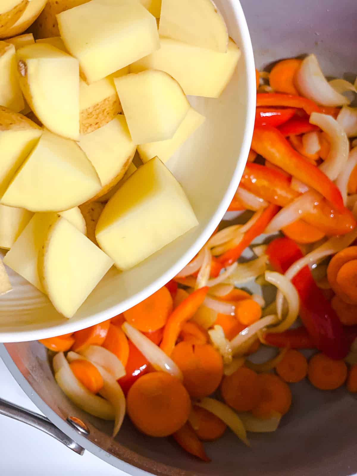 Adding potatoes to a pot with onion, oil, bell peppers, and carrots.