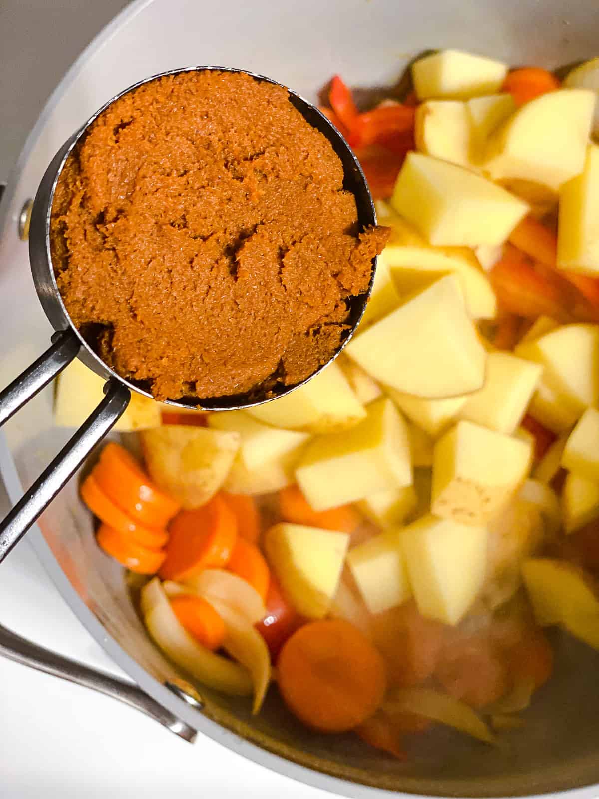 Adding curry paste to a pot with mixed vegetables and oil.