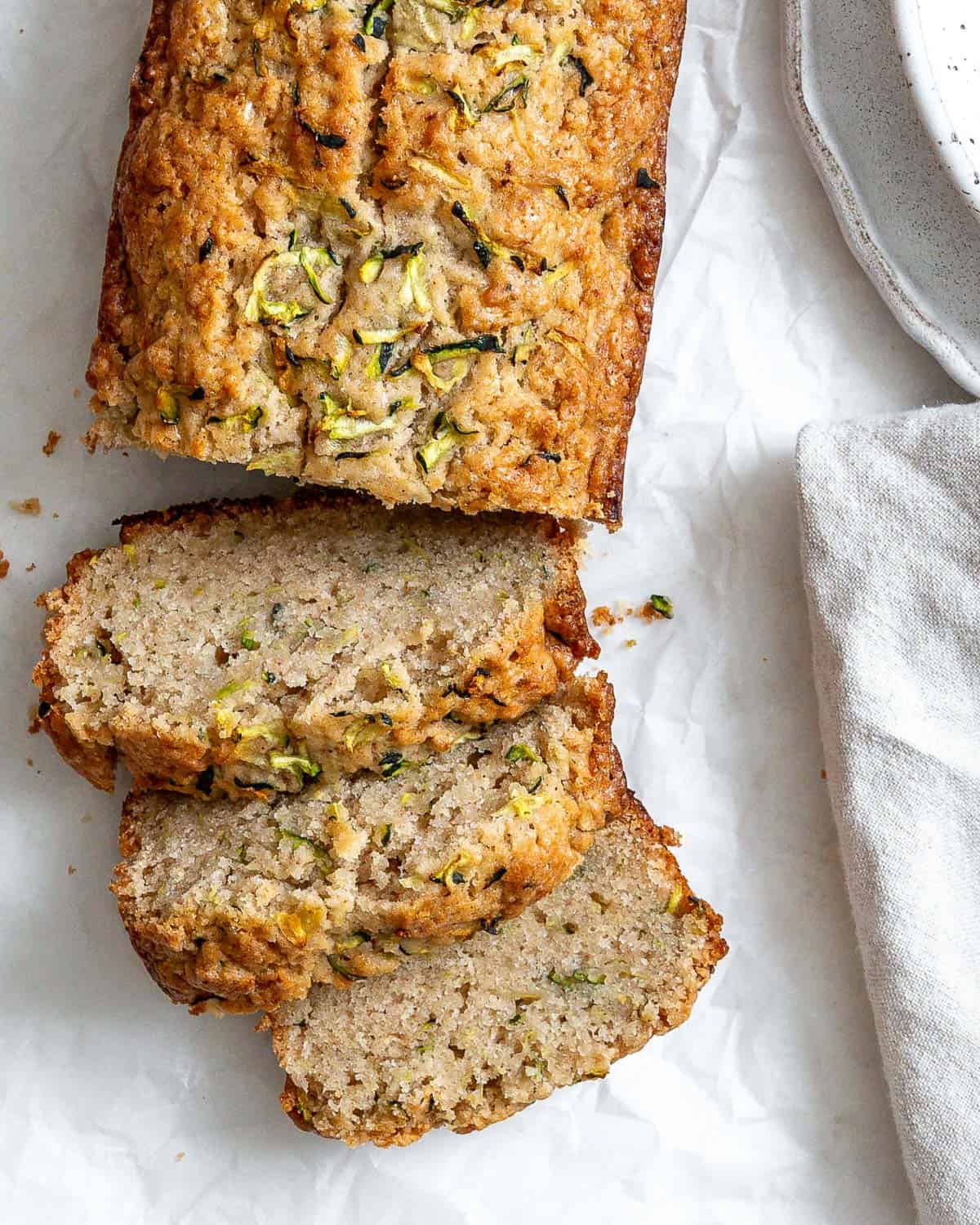 Slices of zucchini bread on white crinkled parchment paper. There's part of a speckled bowl in the top right corner, and part of a gray napkin in the bottom right corner.