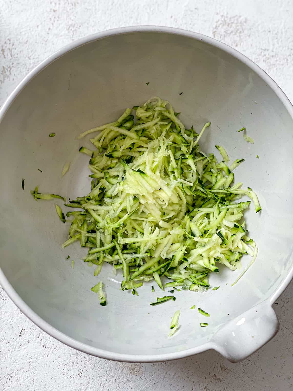 Grated zucchini in a white large bowl on a white textured surface.