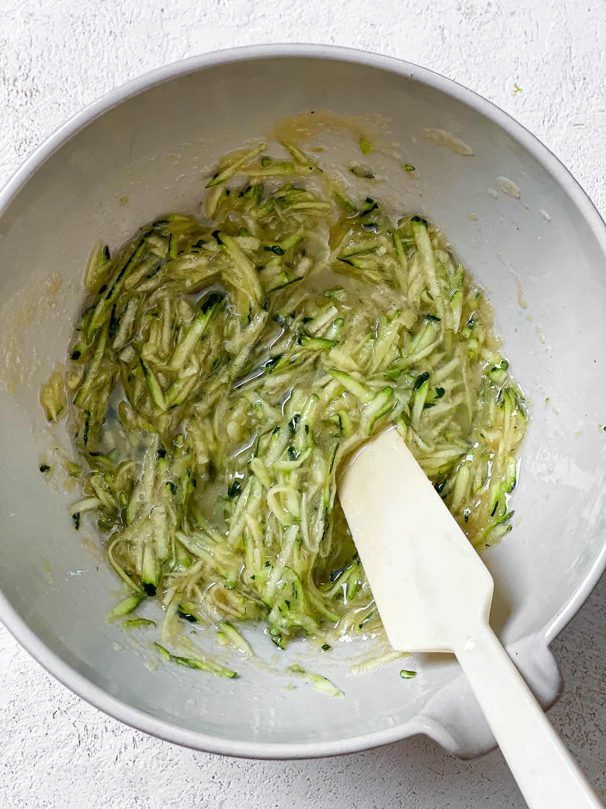 Grated zucchini and wet ingredients in a white bowl with white spatula on a white textured surface.