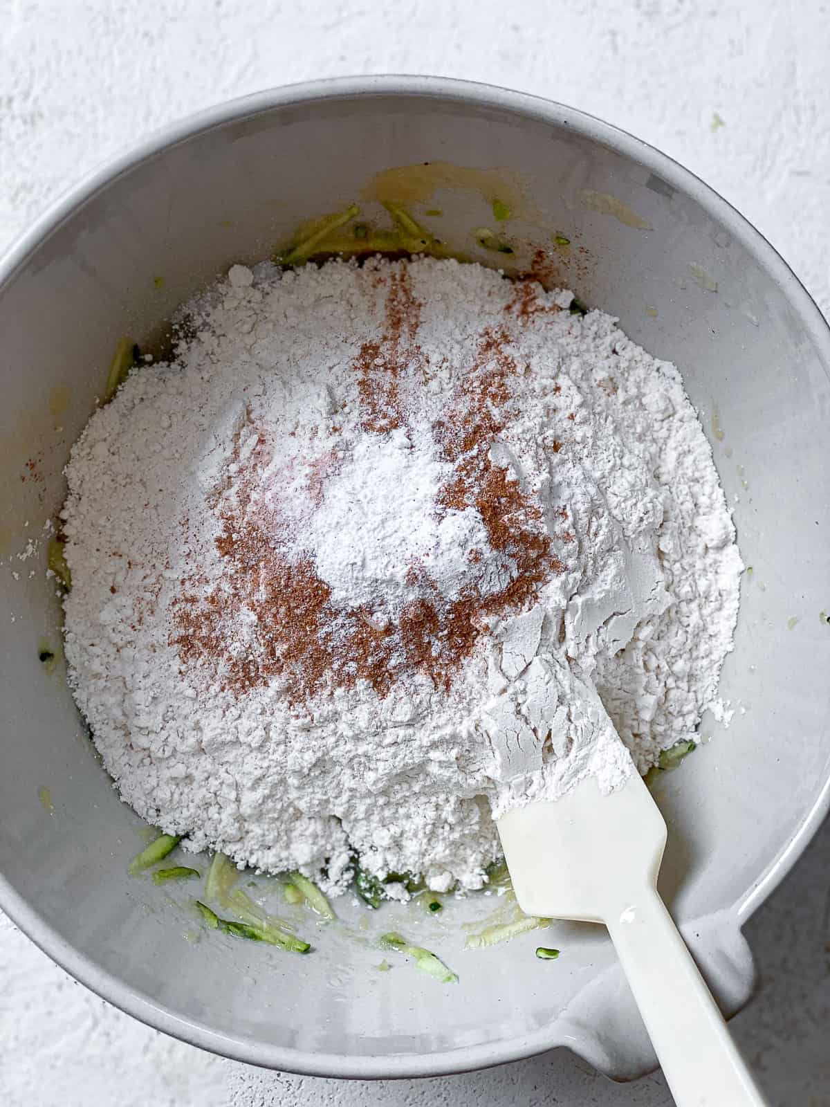 All the ingredients for zucchini bread in a white bowl with white spatula over a textured white surface.