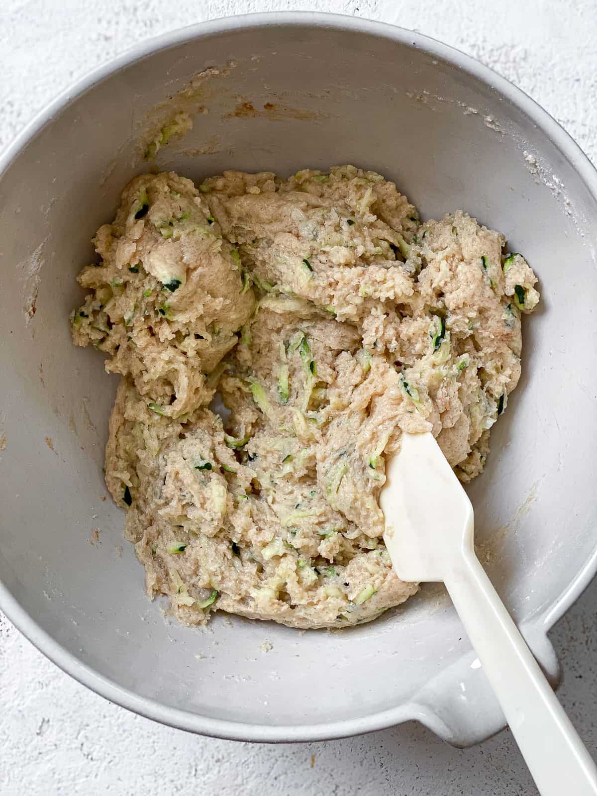 White bowl with all the zucchini bread ingredients mixed together. There's a white spatula in the batter, and the bowl is on top of a white textured surface.