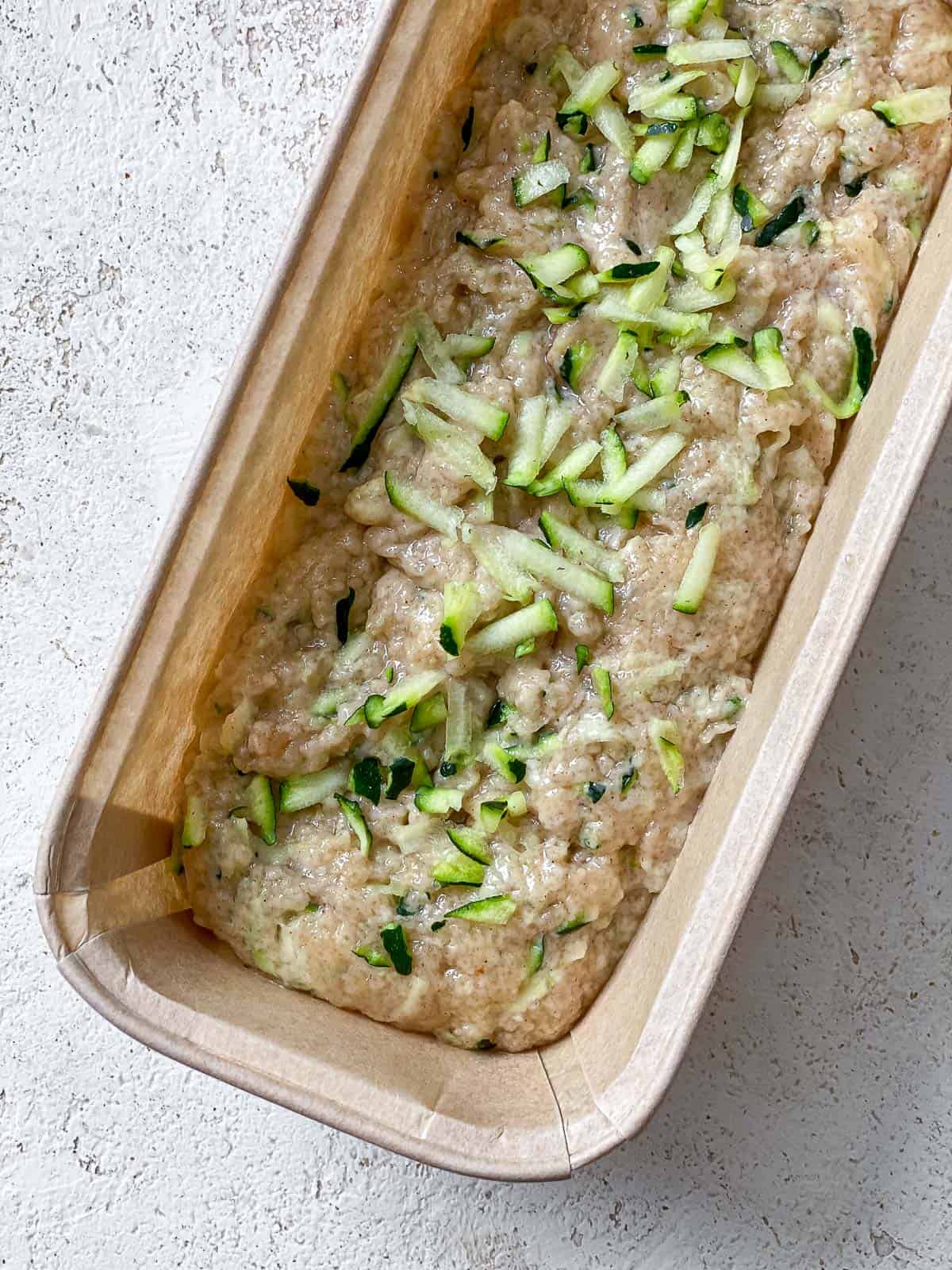 Brown bread loaf pan with zucchini bread batter filled in it sitting on top of a white speckled surface.