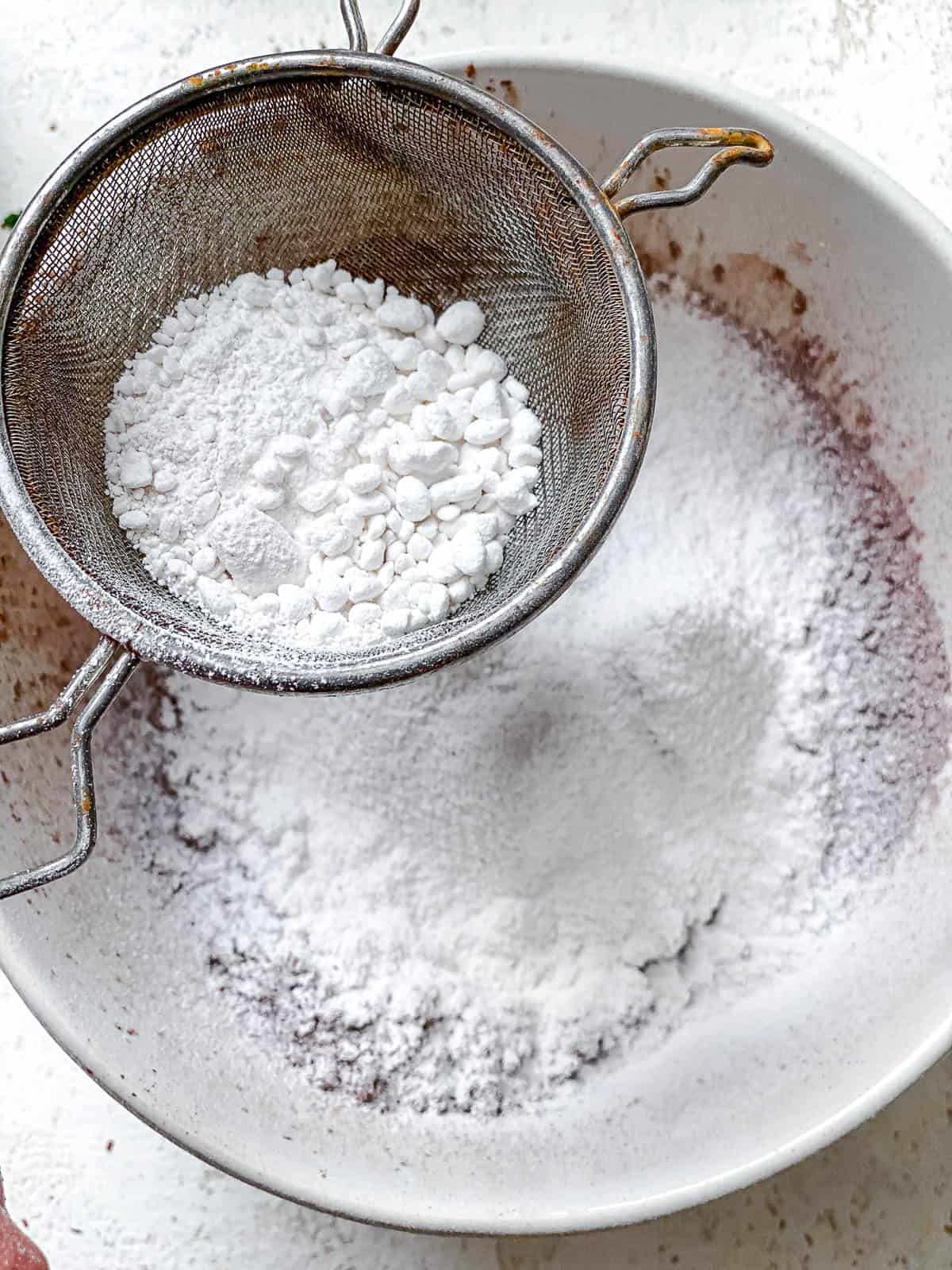 Powdered sugar being sifted through a strainer into cocoa powder in a white bowl