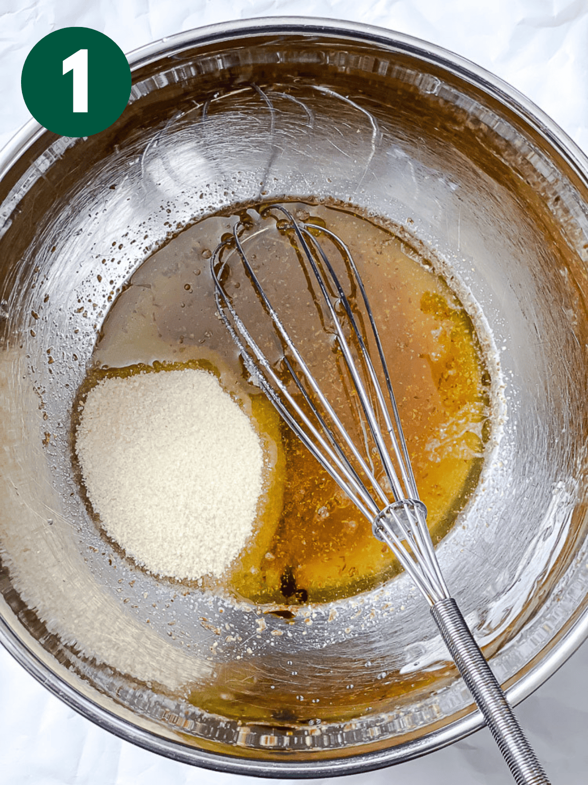 whisking sugar into a golden liquid mixture in a large metal bowl.