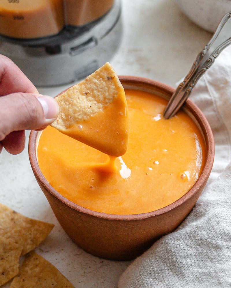 hand dipping a tortilla chip into a small bowl of vegan nacho cheese sauce.