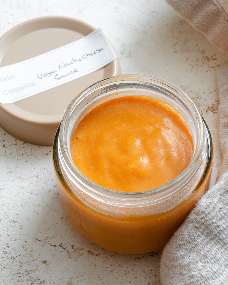 a glass jar filled with vegan nacho cheese sauce with a lid on the side labeled, "vegan nacho cheese sauce".