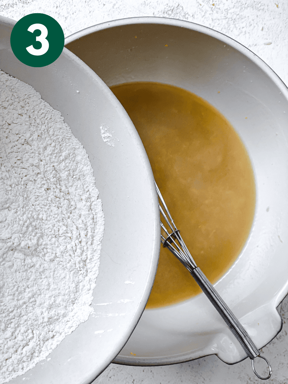 pouring flour into a wet cake batter mixture in a large white bowl.