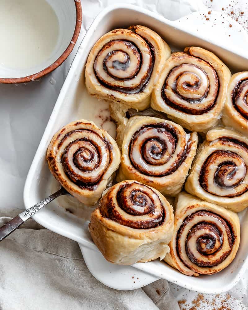 using a metal spoon to lift baked vegan cinnamon rolls out of a white baking pan.