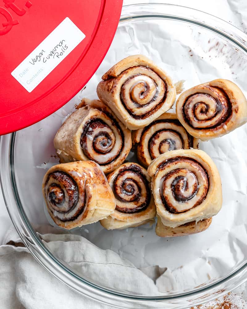 cinnamon rolls in a large glass bowl with a lid on the side labelled "vegan cinnamon rolls".