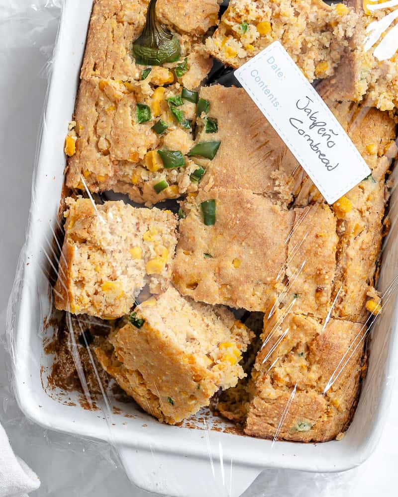 slices of baked jalapeno cornbread in a white baking dish covered in plastic wrap.