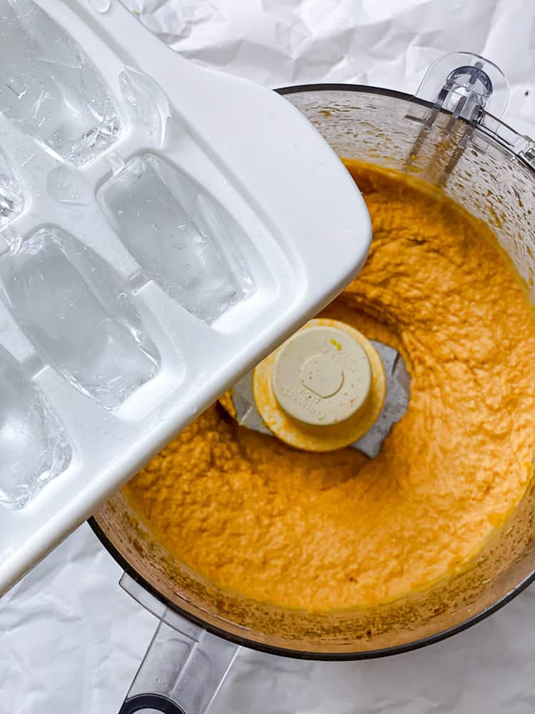 adding ice cubes to blended sweet potato hummus in a food processor.