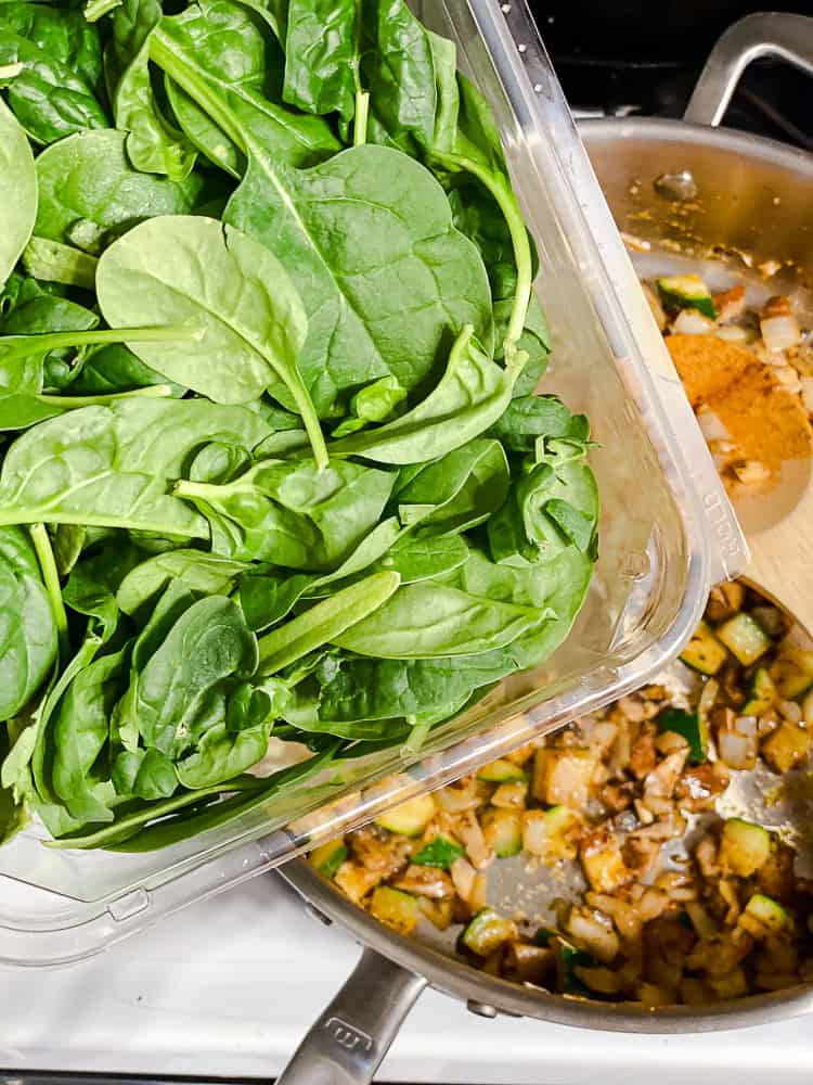 pouring spinach leaves into a skillet full of cooked vegetables.