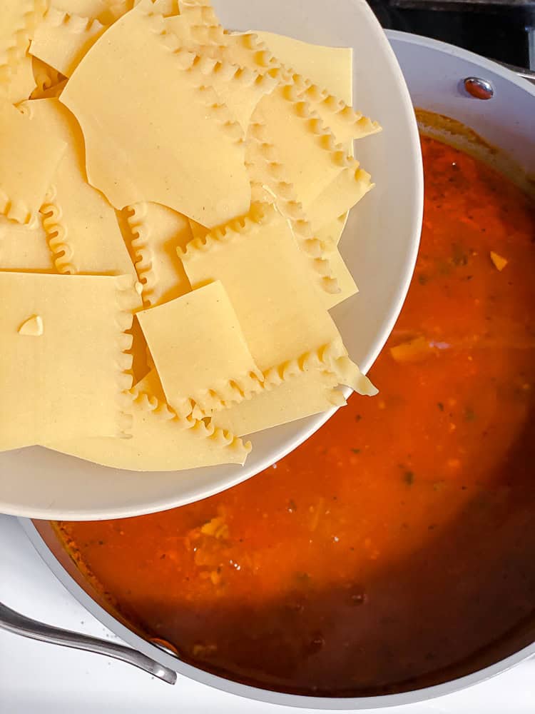 pouring a bowl of lasagna noodles into a pot of vegan lasagna soup.