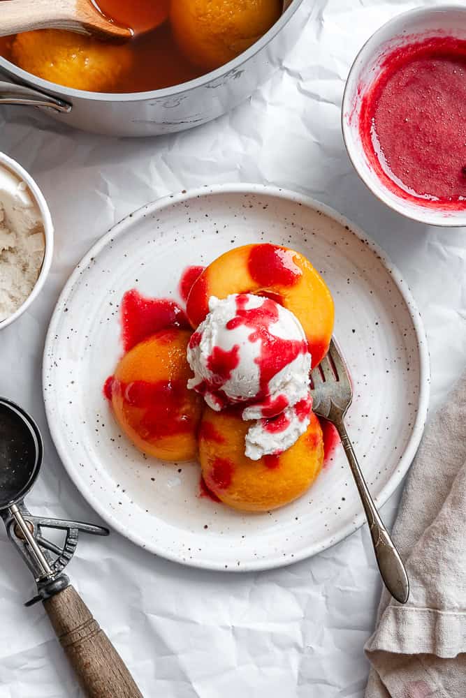 poached peaches topped with raspberry sauce and ice cream on a plate.
