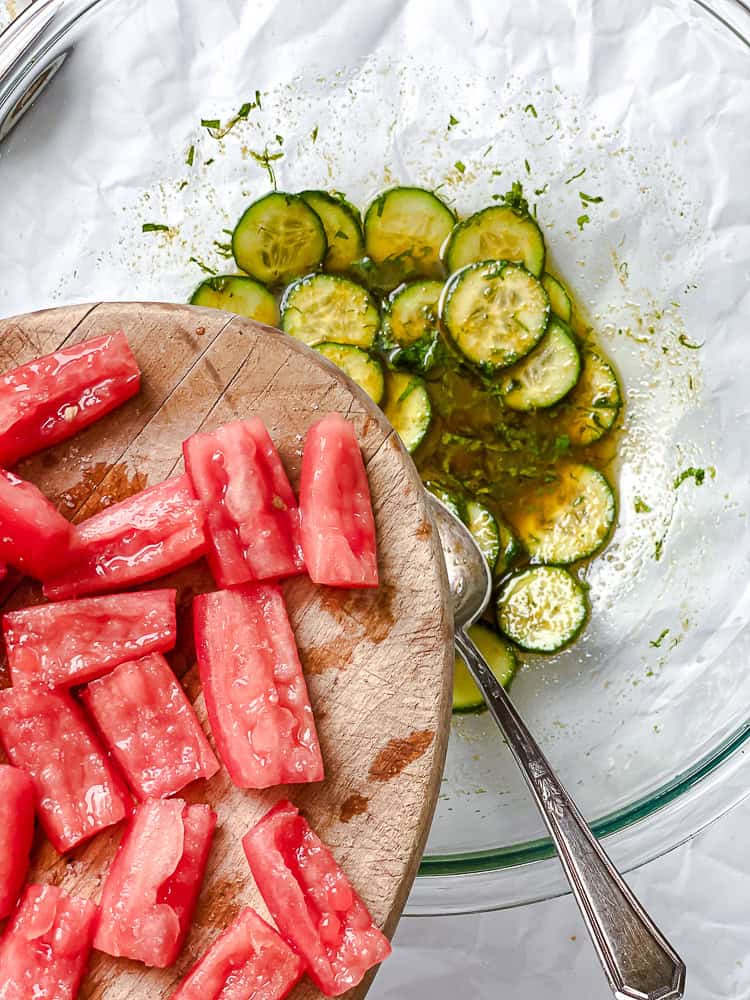 dumping tomatoes into a large glass bowl with marinated cucumbers.