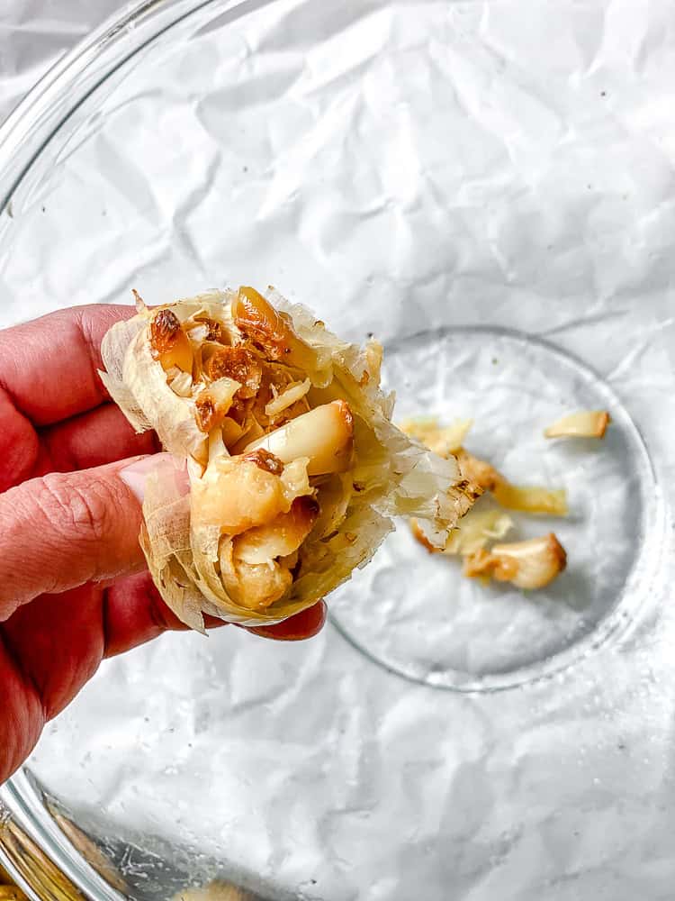 Soft roasted garlic cloves being squeezed out of the head into a bowl.
